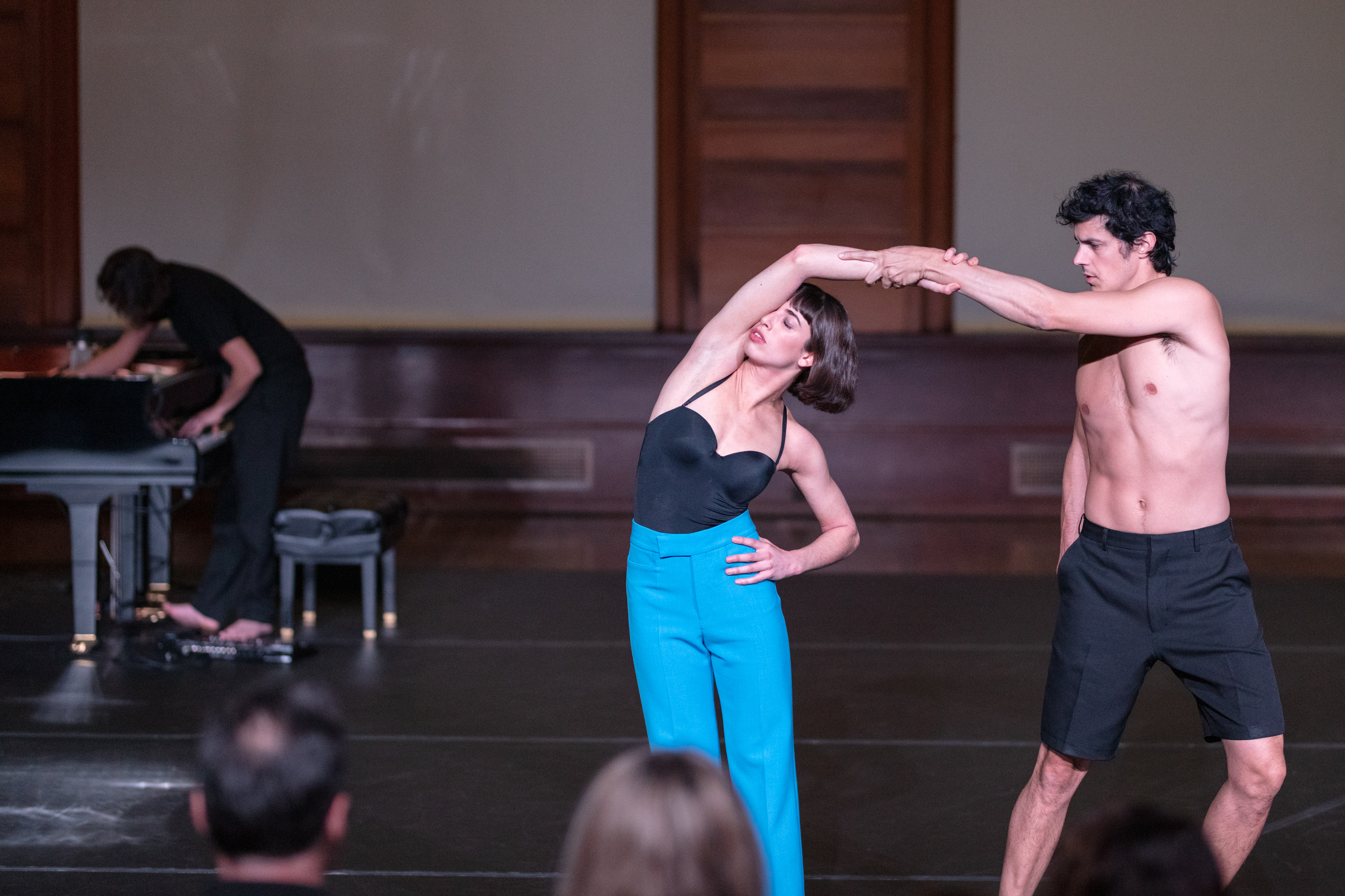 A woman wearing a black tank top and blue pants dances with a man wearing dark shorts, while a man plays piano in the background.