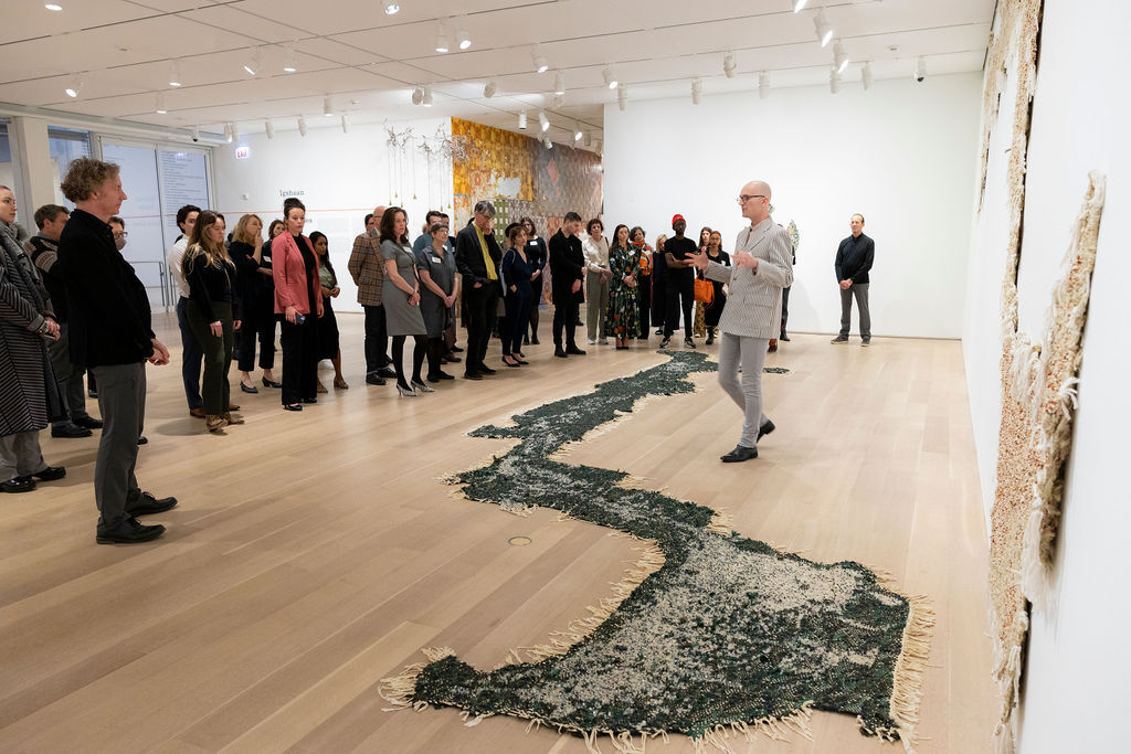 People stand around viewing an artwork made of beads installed on a wooden floor.