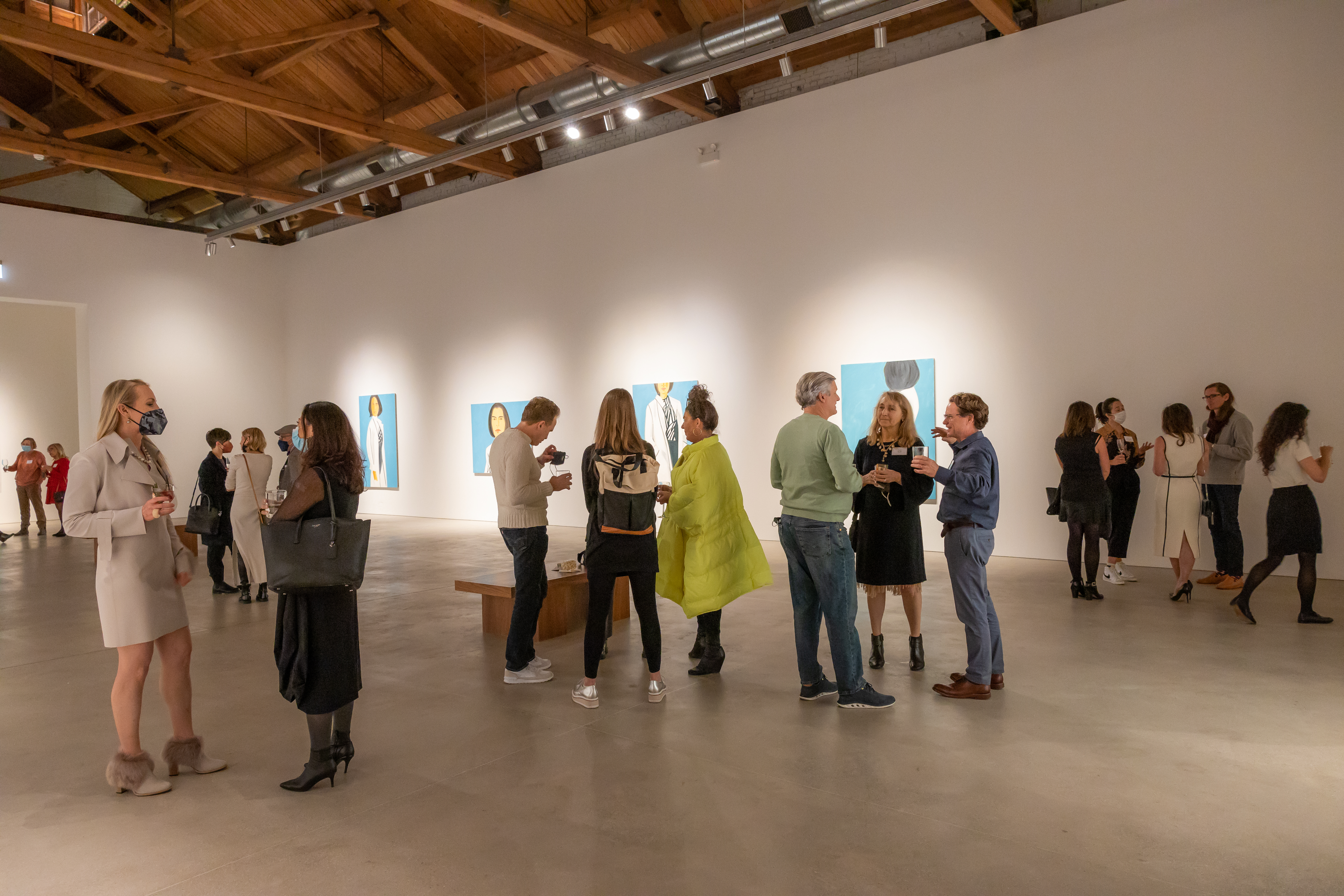 People stand around talking in the middle of a white gallery space with paintings on the walls.